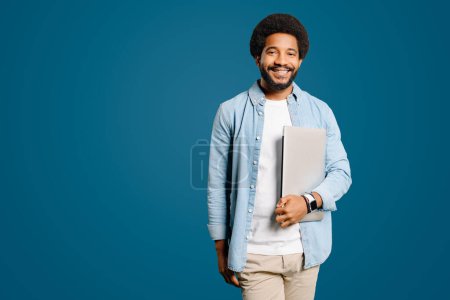 Ein intelligenter und freundlicher junger Mann, der einen Laptop in die Kamera hält, behält sein einnehmendes Lächeln vor dem leuchtend blauen Hintergrund bei und verkörpert den dynamischen Geist jugendlichen Unternehmertums