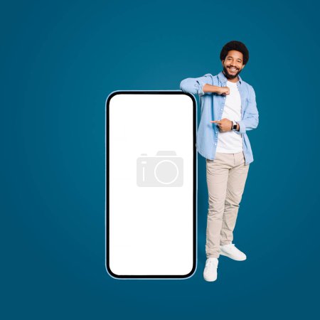 A young Brazilian man with a friendly smile stands beside a blank oversized smartphone, pointing at the potential place for advertising a new app or deal, male freelancer in denim shirt presenting