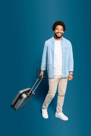 Amical homme brésilien marche avec un pas léger et une expression joyeuse, valise dans la remorque, reflétant le frisson du voyage et de nouvelles découvertes, isolé sur le bleu. Concept de voyage sans soucis