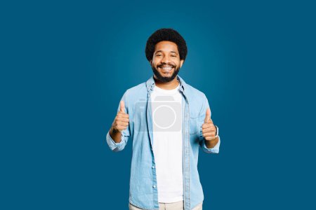With a bright smile, the African-American man gives two thumbs up, conveying double the approval and a cheerful disposition against a vibrant blue background, recommending and agreeing