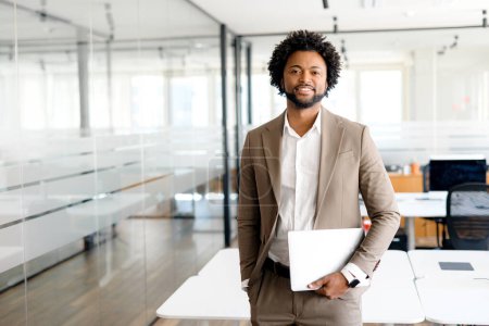 Photo for A cheerful entrepreneur holds his laptop while standing, his posture exuding confidence and professionalism within a sleek office space. Mobility in business, cloud computing, on-the-go professional - Royalty Free Image