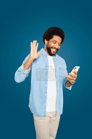 Handsome cheerful Brazilian young man with an afro hairstyle waving while holding his smartphone, portraying a friendly digital interaction or virtual greeting, involved video call or streaming online