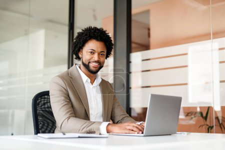 Selbstbewusst und nahbar sitzt ein afroamerikanischer Geschäftsmann an seinem Schreibtisch in einem modernen Büro, die Hände auf der Laptop-Tastatur, bereit, die Herausforderungen der heutigen Zeit anzugehen.