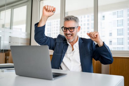 Overjoyed 60s mature businessman is gesturing victory with his arms raising up, senior manager scream with triumph, winning in game, having received great news of a good deal