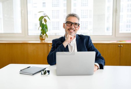 Gut aussehender, reifer Geschäftsmann mit Laptop, der in einem modernen Büro am Arbeitsplatz sitzt, selbstbewusster Senior-Manager der 50er Jahre, der in die Kamera schaut und lächelt