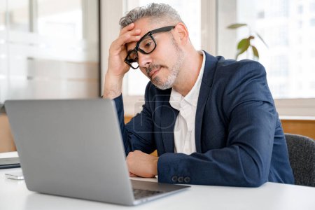 Téléchargez les photos : Homme d'affaires mature fatigué utilisant un ordinateur portable pour travailler, homme en tenue formelle se sentant fatigué et épuisé, assis au bureau dans un bureau moderne, doutant et pensant - en image libre de droit