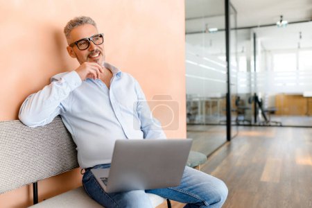 With a smile and a contemplative look, a businessman works on his laptop in a relaxed setting, embodying the blending of comfort and corporate acumen