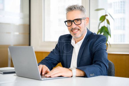 Freundlicher und positiver reifer männlicher Angestellter oder Manager mit Laptop, leitender Geschäftsmann in formeller Kleidung und Brille, der auf E-Mails im modernen Büro reagiert