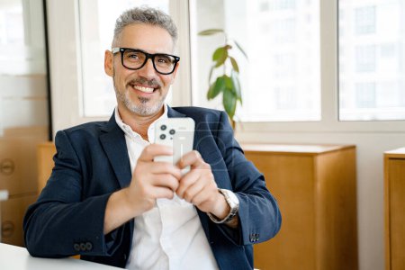 A seasoned executive engages with his smartphone, his radiant smile suggesting a successful business communication in a modern office space