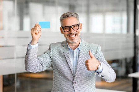 Photo for A stylish mature businessman holds up a blank business card, signifying readiness for networking and professional exchanges. His thumbs-up gesture indicates approval and a positive business approach. - Royalty Free Image