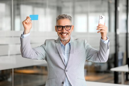 Cheerful happy mature businessman holding smartphone and credit card, received money transfer, salary, income from investments, benefits standing with the office background, online shopping, e-banking