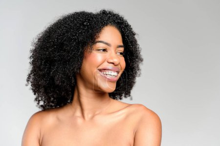 A joyful African-American woman beams with a captivating smile, her natural beauty accentuated by her curly hair against a plain background. The shot perfect for beauty and lifestyle themes.