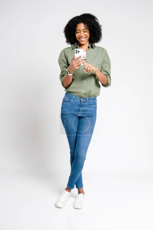 Femme afro-américaine au sourire captivant, profondément absorbée par son smartphone, mettant en valeur la joie de la connectivité numérique, elle incarne parfaitement un mode de vie urbain détendu mais à la mode.