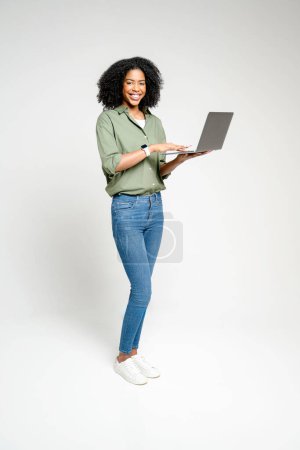 Capturée dans un plan complet, une femme afro-américaine avec une expression joyeuse utilisant l'ordinateur portable, mettant en valeur la polyvalence des appareils sans fil modernes qui permettent la mobilité et la flexibilité