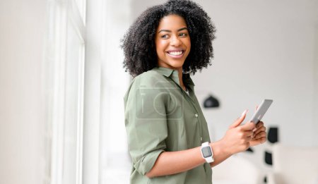 Brillante de felicidad, una mujer afroamericana sostiene y opera una tableta, su compromiso con el dispositivo que sugiere una experiencia en línea agradable en un ambiente interior cómodo y brillante