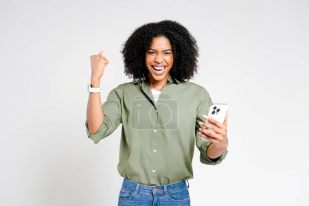 Foto de Una mujer afro-americana emocionada con una sonrisa dentada celebra una victoria en su teléfono inteligente, su triunfo de señalización de puño y euforia. - Imagen libre de derechos