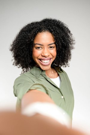 Con una alegre postura de selfie, una mujer afroamericana captura su propia imagen, su radiante sonrisa sugiriendo un momento de celebración personal o una actualización de las redes sociales en un entorno profesional