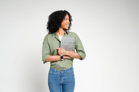 Woman stands with a tablet in hand, playfully looking away as if in thought, against white backdrop. Casual yet contemplative scene, ideal for illustrating ideas around learning or digital exploration