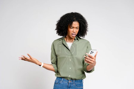 Téléchargez les photos : Surprise et légèrement perplexe, cette femme afro-américaine regarde son écran de smartphone, son expression mêlant confusion et curiosité, sur fond blanc minimaliste. - en image libre de droit