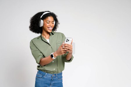 Téléchargez les photos : Femme afro-américaine dans les écouteurs à la mode sourit tout en utilisant son smartphone, choisir une chanson ou bavarder avec des amis, soulignant l'intégration transparente de la technologie dans les moments quotidiens - en image libre de droit