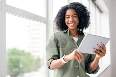Curly friendly African-American young woman smiles brightly near a window, her interaction with the tablet suggesting an enjoyable work-from-home experience.
