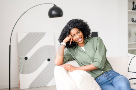 Téléchargez les photos : Avec une inclinaison ludique de la tête et un sourire charmant, une femme afro-américaine se détend sur un canapé dans une pièce bien éclairée qui respire le style contemporain et le confort, illustrant la vie moderne à la maison. - en image libre de droit