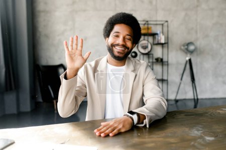 Young businessman waves hello with a warm, inviting smile, positioned in a well-lit office setting that reflects a modern and welcoming workplace