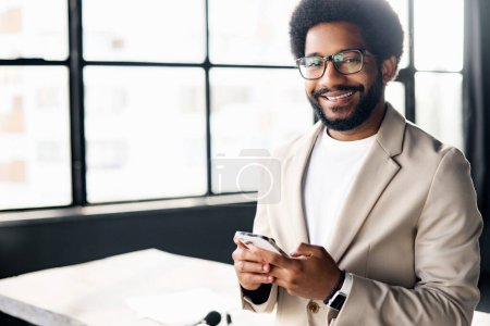 This professional using his smartphone standing in a stylish office setting. The large windows behind him fill the room with light. African-American male employee texting online on the mobile phone