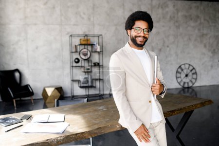 Confident young businessman stands by his workspace, holding a laptop in one hand, his smile exuding confidence and readiness for the challenges of the day. The image epitomizes professional readiness