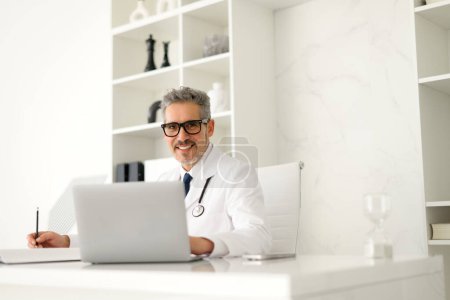 Reifer Arzt mit freundlichem Auftreten tippt während er Notizen macht auf einen Laptop und zeigt das Multitasking, das in modernen datengesteuerten Arztpraxen erforderlich ist