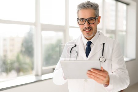 El médico senior con sonrisa confiada y brillante sostiene una tableta en la oficina moderna, refleja la experiencia positiva del paciente que pretende proporcionar. Práctica médica contemporánea