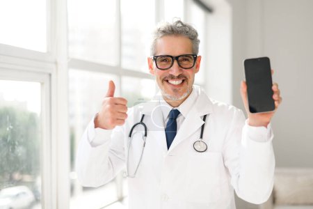 Doktor, mit einem Stethoskop um den Hals, gibt Daumen nach oben, während er ein Telefon mit einem leeren Bildschirm hält, was auf eine erfolgreiche Patienteninteraktion oder einen positiven Gesundheitsbericht hindeutet, der medizinische App darstellt