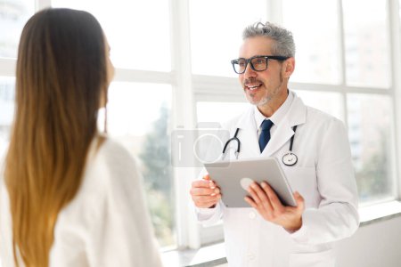 A senior doctor with grey hair and glasses is holding a digital tablet and talking with a young female patient, in a clinic office that offers a panoramic city view. Technology in healthcare
