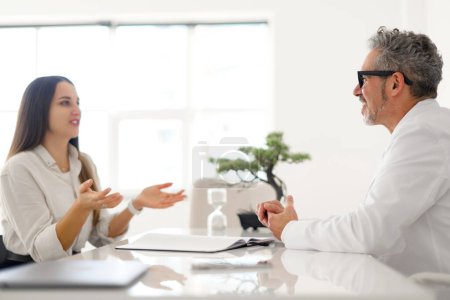 Photo for A senior male doctor with grey hair, wearing glasses and a white lab coat, attentively listens to a young female patient. Communicative and professional nature of the medical consultation - Royalty Free Image
