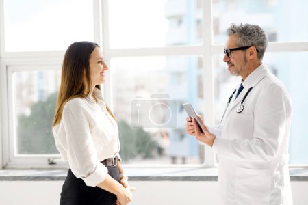 Ein leitender Arzt mit einem Hauch von Grau im Haar lächelt, während er eine Tablette in der Hand hält und unbeschwert mit einer jungen Patientin in einer Klinik mit hellem Stadtbild draußen interagiert.