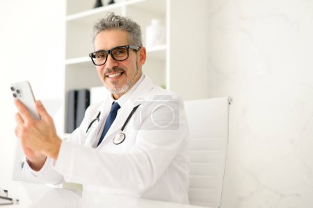 A senior doctor with grey hair is pictured in a well-lit clinic, holding a smartphone, staying connected with patients or the use of health apps in medical consultations.