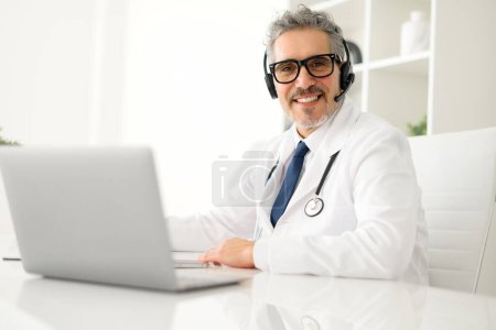 Mature grey-haired doctor wearing headset and white coat using a laptop, giving a reassuring smile during an online consultation, indicating a comfortable and professional telemedicine experience.