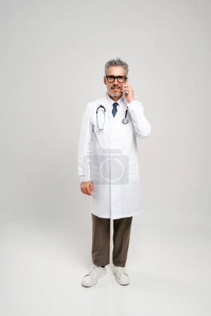 The grey-haired doctor stands upright while talking on the phone, reflecting the active role of physicians in coordinating patient care even from a distance.