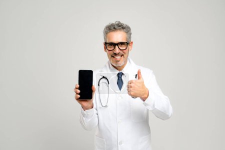 Fröhlicher Oberarzt, der den Daumen hebt, während er ein Smartphone hält, eine medizinische Anwendung unterstützt oder eine erfolgreiche Patientenkommunikation bestätigt, fördert Vertrauen und Positivität im Gesundheitswesen