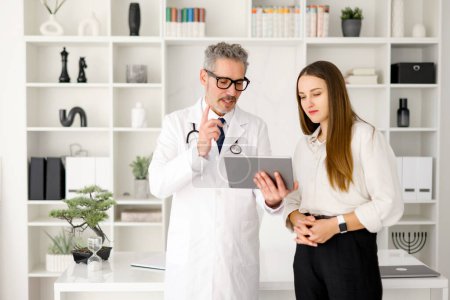 Téléchargez les photos : Un médecin homme aux cheveux gris en manteau blanc discute des informations médicales avec une jeune femme, à l'aide d'une tablette pour améliorer la compréhension, debout dans un bureau moderne - en image libre de droit
