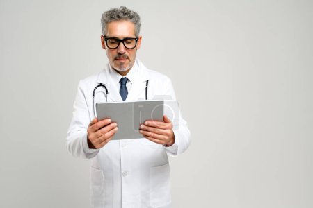 Senior doctor with grey hair is deeply focused on tablet, reviewing patient information or medical records. His intense concentration demonstrates integration of technology in modern medical practice