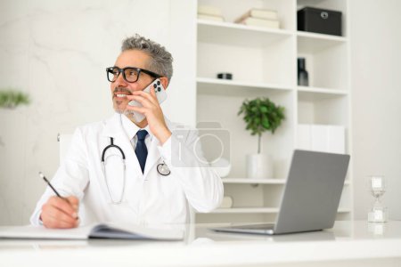 Cheerful senior doctor engaging in a phone conversation, pen in hand, ready to note down important patient information, set in a neat and professional clinic environment.