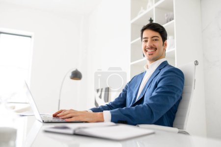 Photo for An affable Hispanic businessman smiles, looks at the camera while using a laptop in a well-lit, stylish office, representing a positive and successful business attitude. Male employee in the workplace - Royalty Free Image