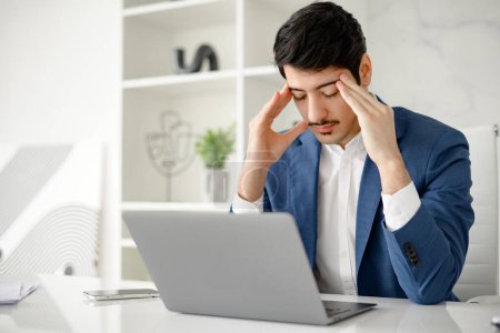 A Hispanic businessman shows signs of stress, massaging his temples while focusing on his laptop in an office space, illustrating the demanding aspects of business and the need for problem-solving.