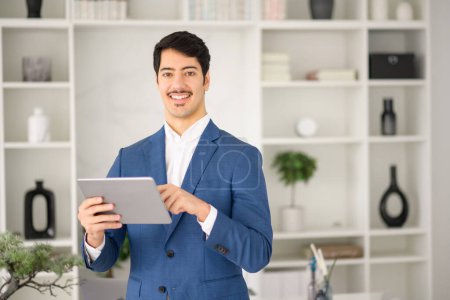 A professional Hispanic businessman in a sharp blue suit confidently uses a tablet in his office, exemplifying the modern blend of style and technology in business