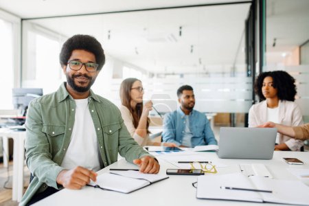 Ein selbstbewusster Mann in lässigem grünem Hemd und Brille lächelt in die Kamera, umgeben von fokussierten Kollegen in einem modernen Büroumfeld, das eine kollaborative und integrative Arbeitsumgebung suggeriert.