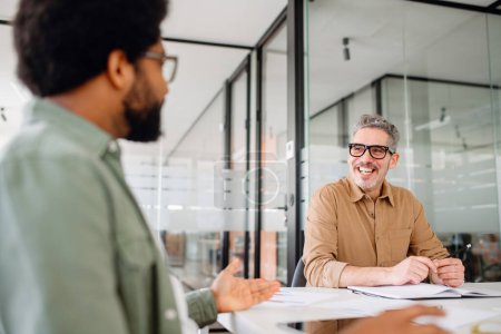 Zwei männliche Berufskollegen führen im Büro ein freundliches Gespräch, der ältere Mann mit Brille zeigt ein warmes Lächeln, das auf ein positives Arbeitsumfeld hinweist.