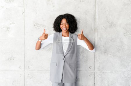 A beaming African-American businesswoman gives a double thumbs up, signaling approval and success, set against a plain gray background conveying simplicity and focus.