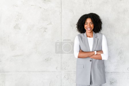 With arms crossed and a confident stance, this professional African-American woman exudes strength and competence against a textured backdrop, embodying the modern corporate leader.