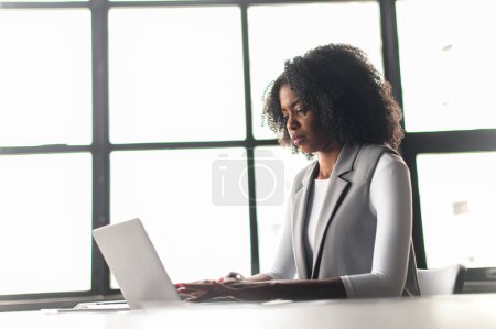 Téléchargez les photos : Une femme afro-américaine s'assoit et utilise un ordinateur portable, sa silhouette encadrée par les lignes géométriques de grandes fenêtres derrière elle, parfaite pour les thèmes des espaces de travail modernes et des professionnels concentrés. - en image libre de droit